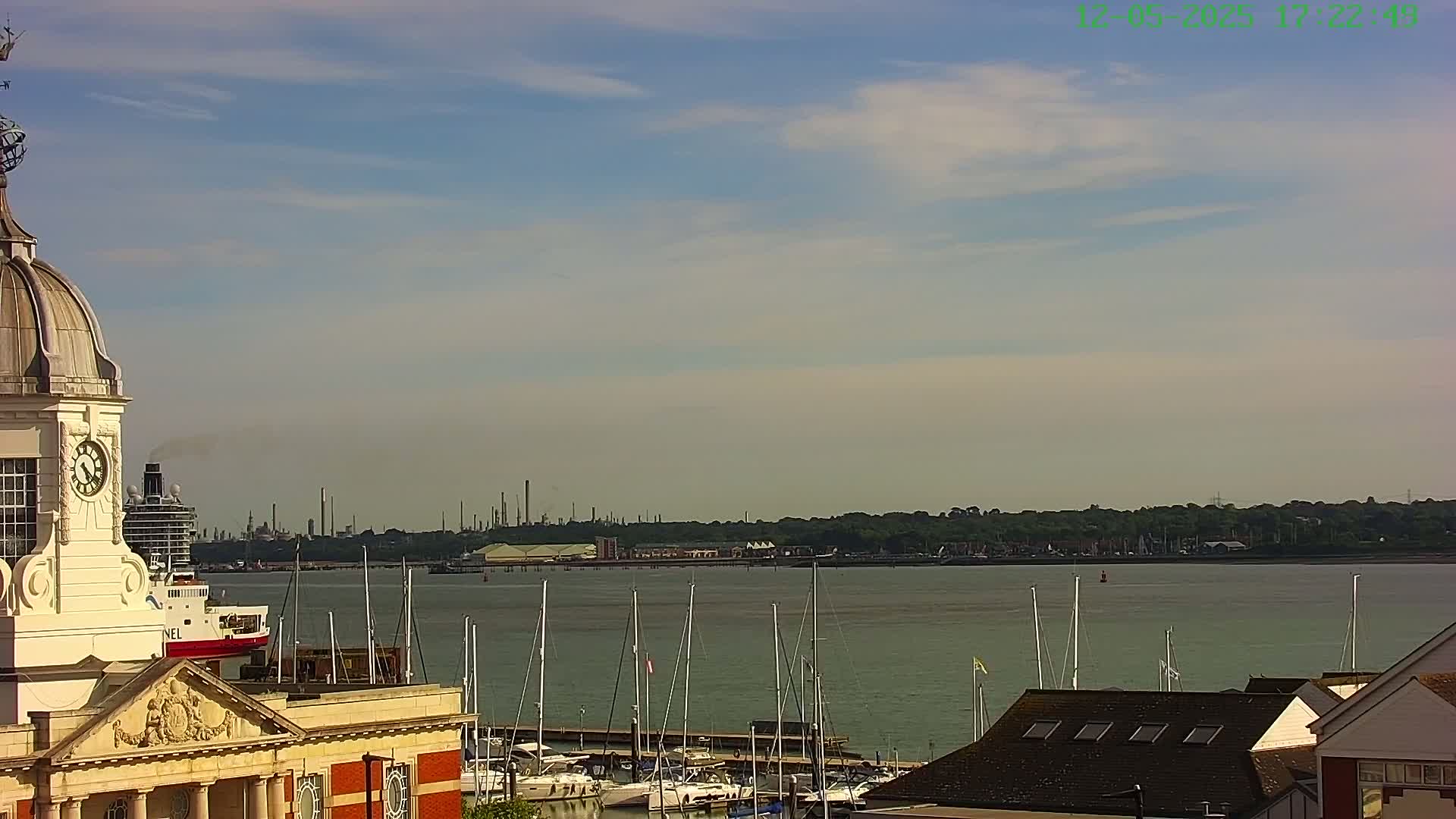 A clock tower overlooks a calm body of water with numerous sailboats docked, and an industrial area visible in the distance under a partly cloudy sky.