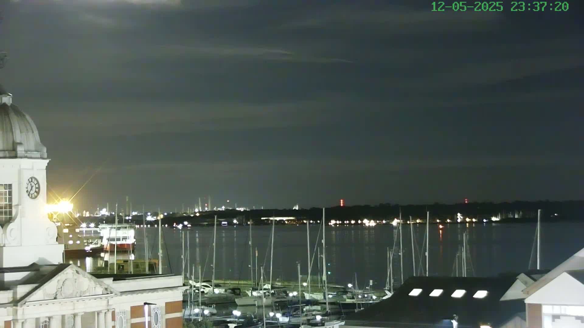 A nighttime view shows a harbor filled with docked sailboats under a dark, cloudy sky, with a building featuring a dome and clock visible in the foreground.