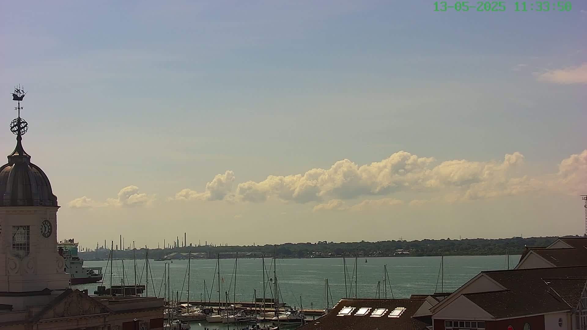 A partly cloudy sky overlooks a harbor filled with sailboats and a distant industrial skyline.