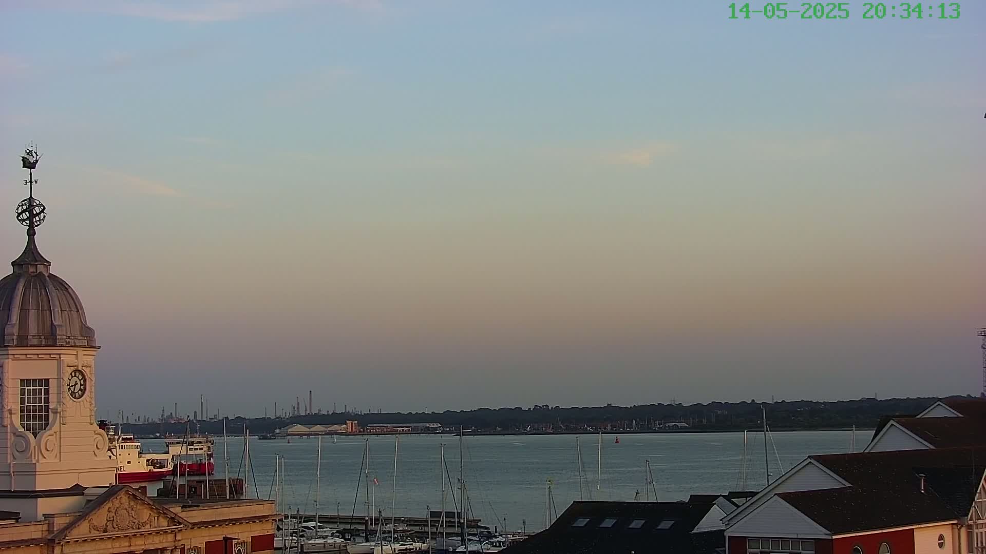 A harbor scene at dusk shows a calm body of water with numerous sailboats and a few ships, viewed from a vantage point overlooking buildings with a clock tower in the foreground.