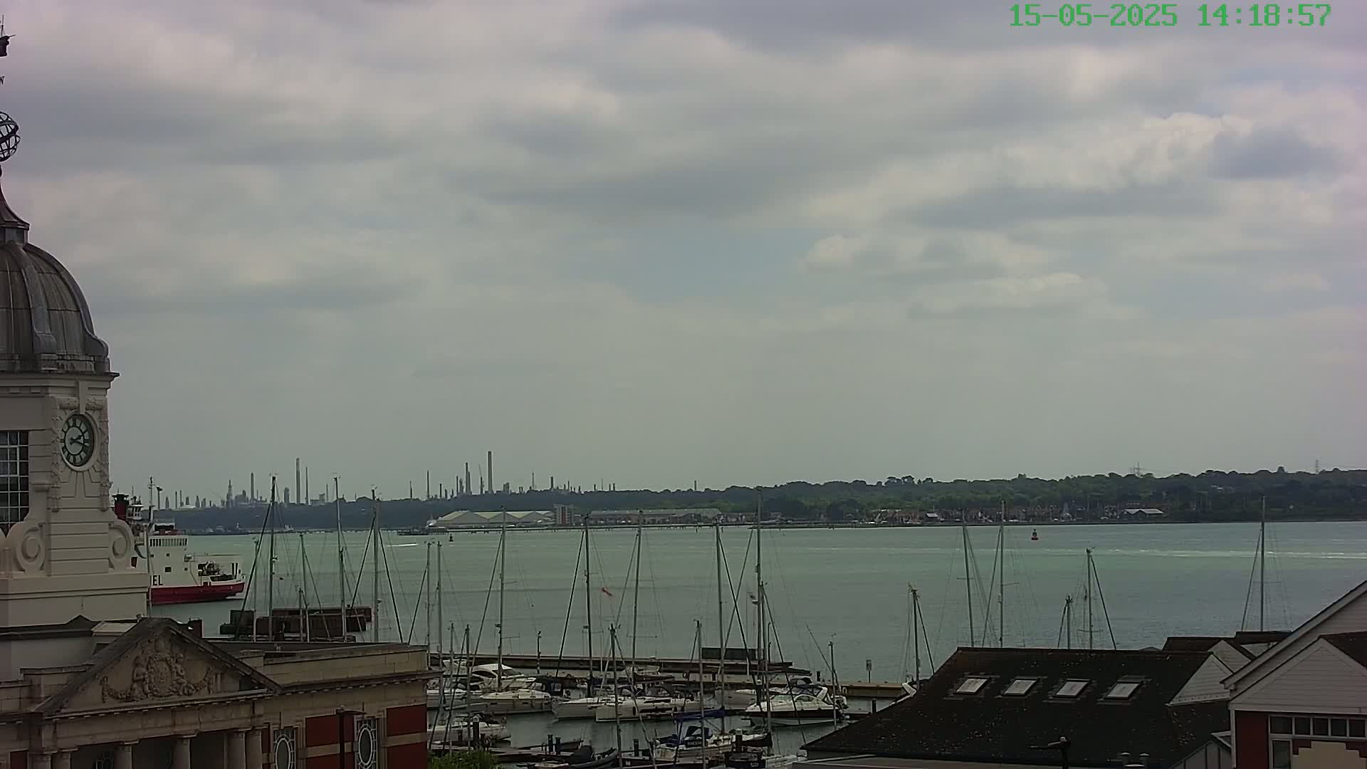 A clock tower overlooks a harbor filled with sailboats, a ferry, and a distant industrial skyline under a cloudy sky.