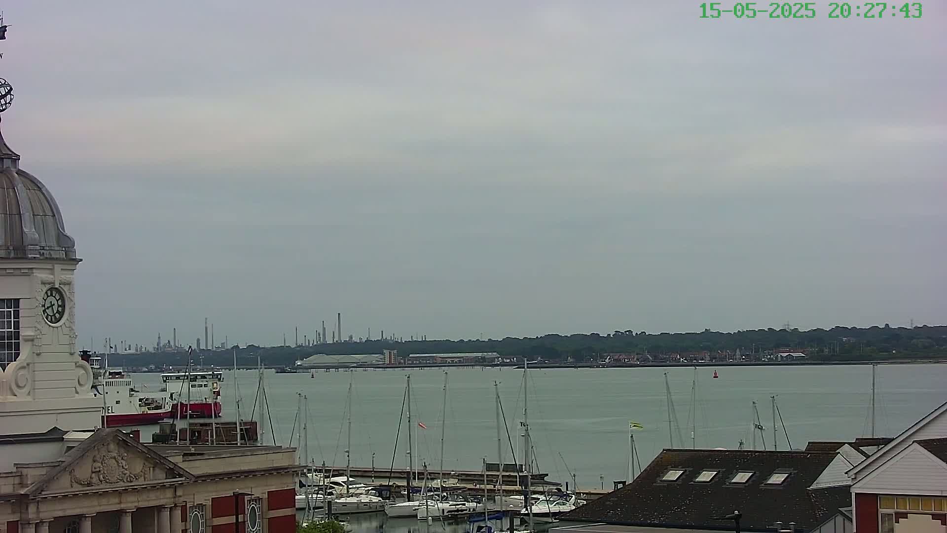 A clock tower overlooks a harbor filled with sailboats on a calm, overcast day, with an industrial skyline visible in the distance.