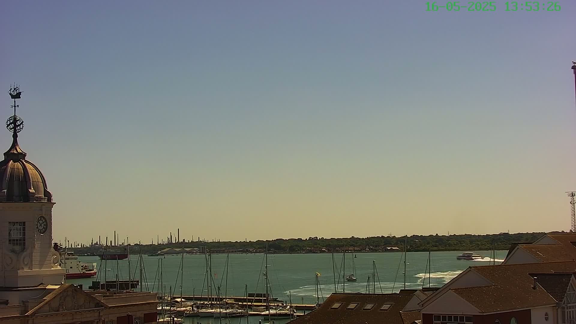 A harbor filled with sailboats is seen from atop a building with a domed clock tower under a clear, sunny sky.
