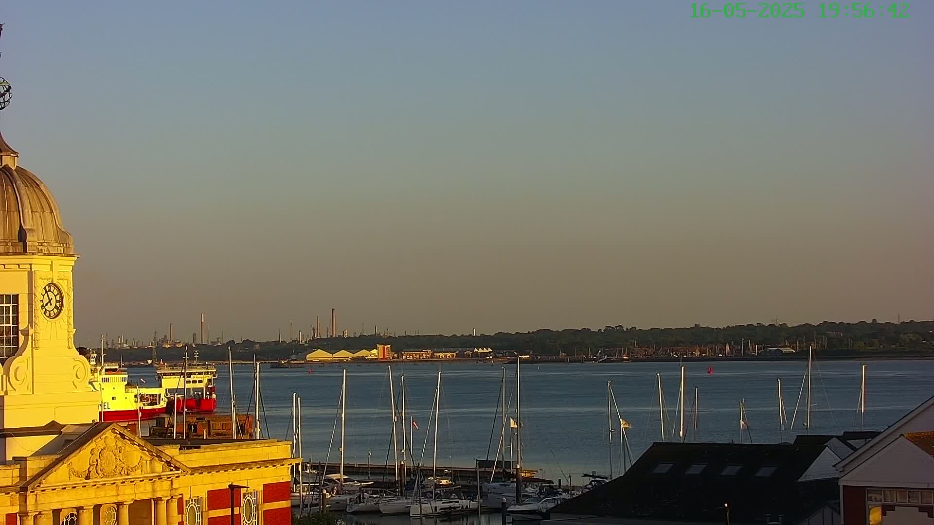 A yellow clock tower overlooks a calm body of water filled with sailboats at sunset, with an industrial landscape visible in the distance.