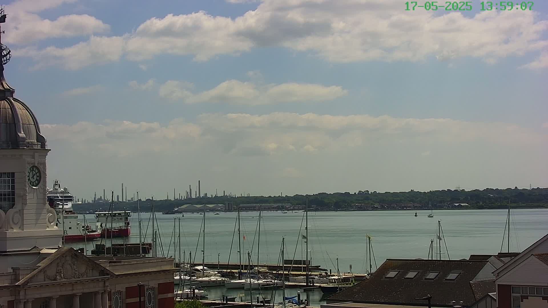 A partly cloudy day shows a harbor filled with sailboats and a docked ship, viewed from a building with a clock tower, and a distant industrial skyline.