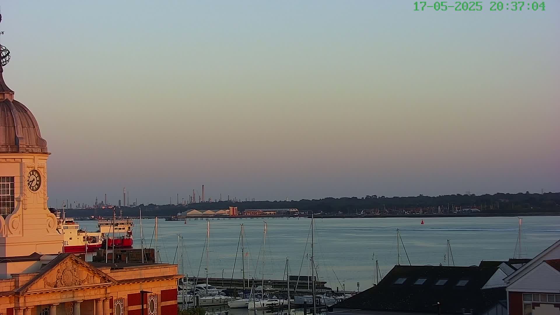 A clock tower overlooks a calm body of water at sunset, with numerous sailboats in a marina and an industrial skyline visible in the distance.