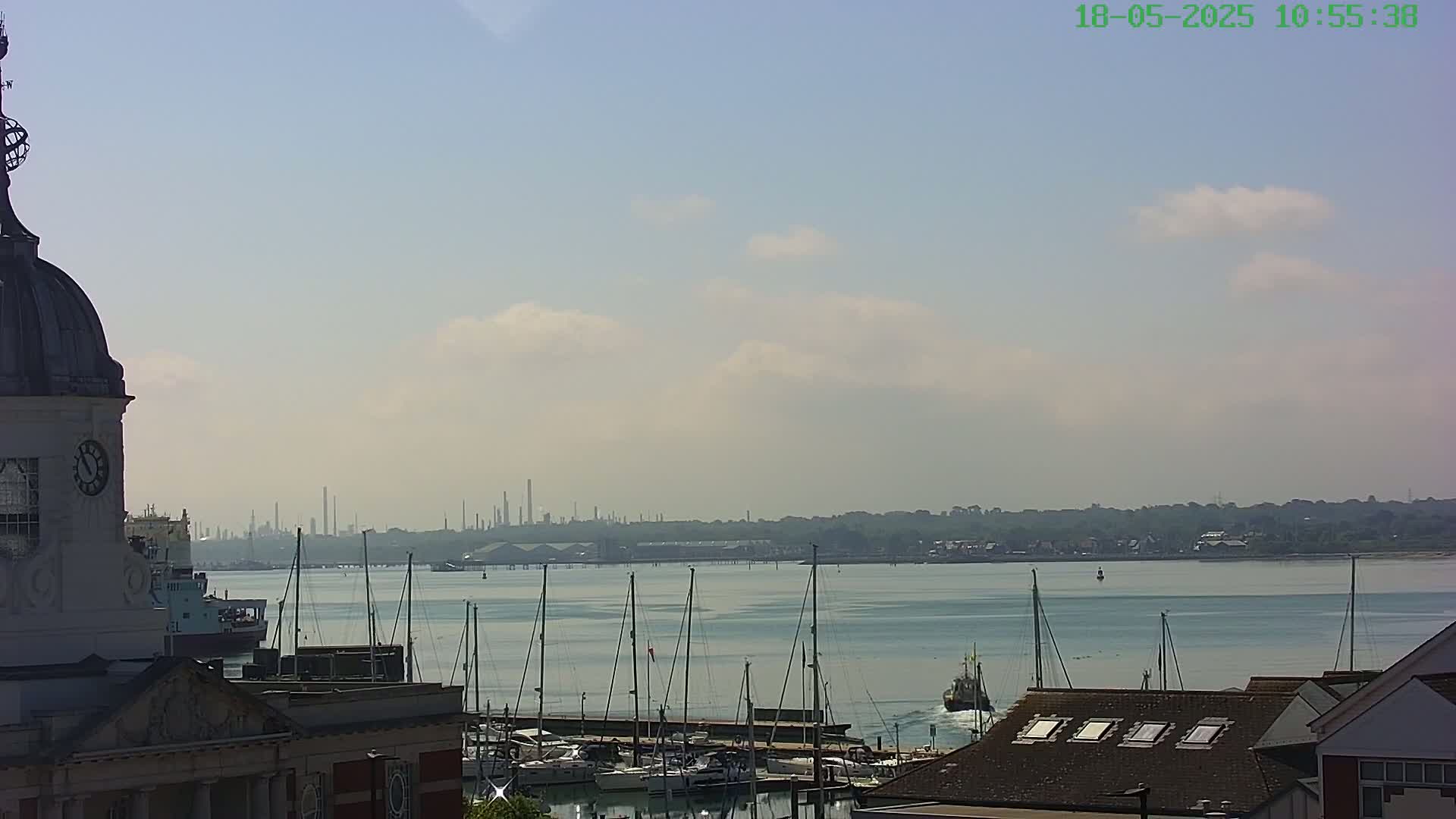 A hazy, sunny day reveals a harbor filled with sailboats and a distant industrial skyline viewed from atop a building with a clock tower.