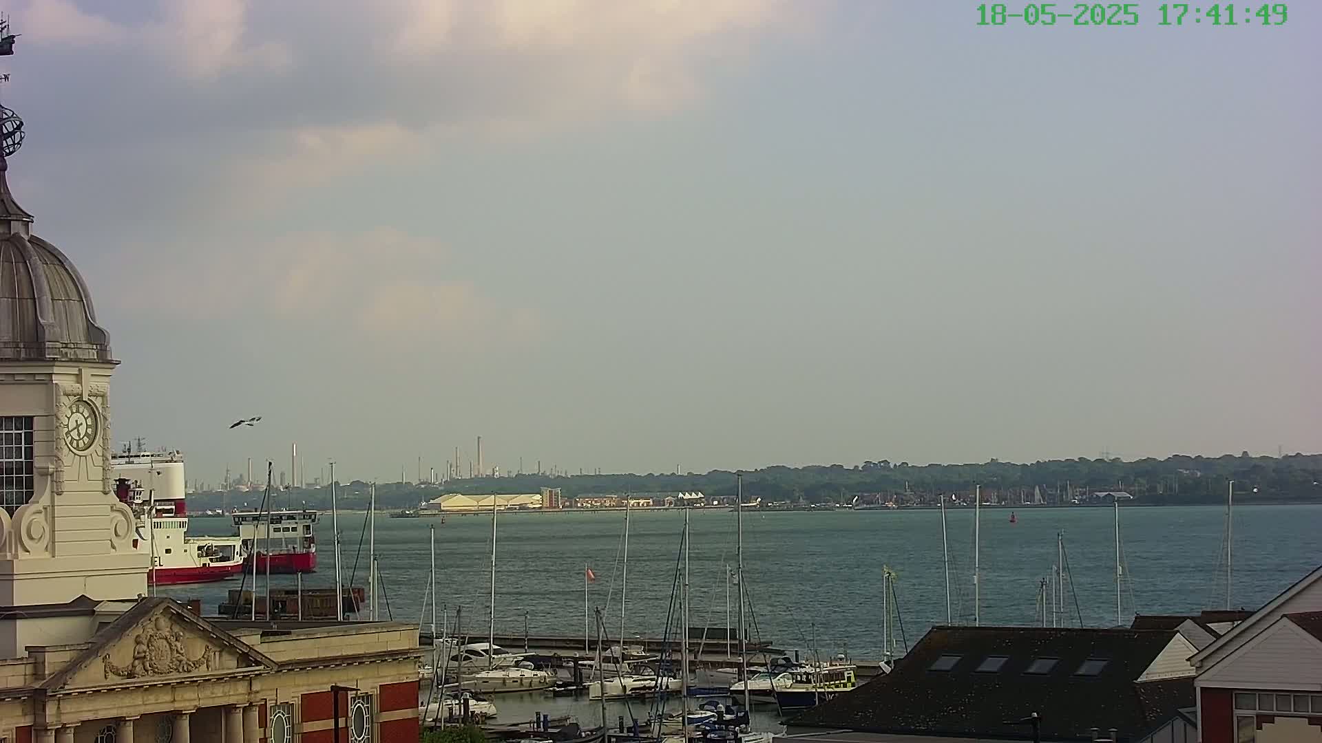 A partially cloudy day shows a harbor filled with sailboats and a larger vessel, viewed from a vantage point above a building with a dome and clock tower, with an industrial area visible in the distance across the water.