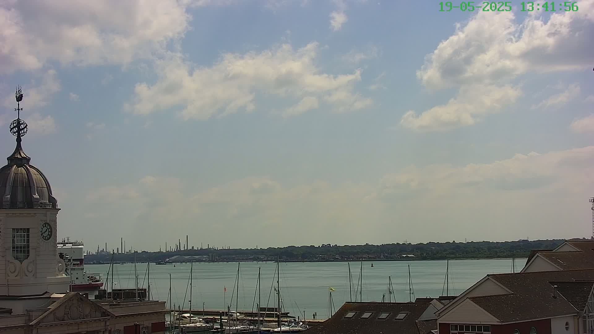 A partly cloudy day reveals a harbor with numerous sailboats, a distant industrial skyline, and a building with a dome and weathervane in the foreground.