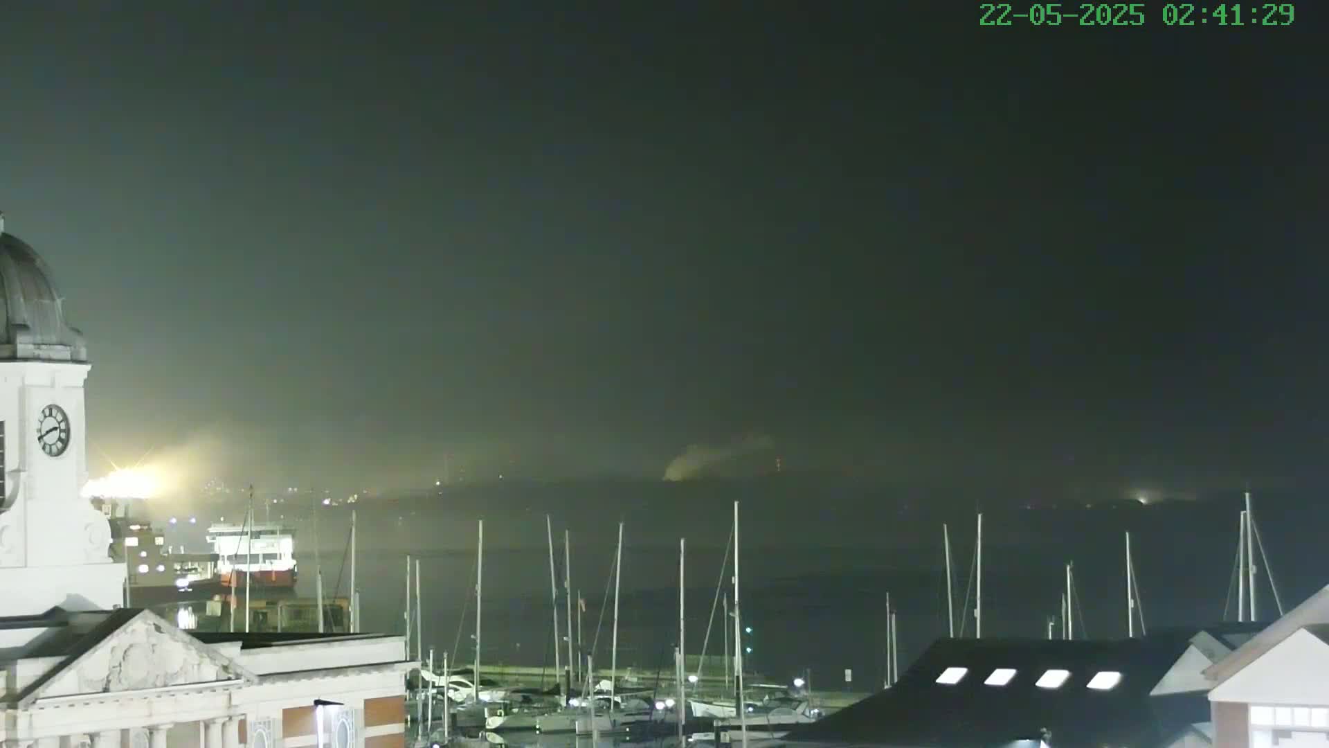 A nighttime view of a harbor filled with docked sailboats, seen from atop a building with a clock tower, under a hazy, dark sky.