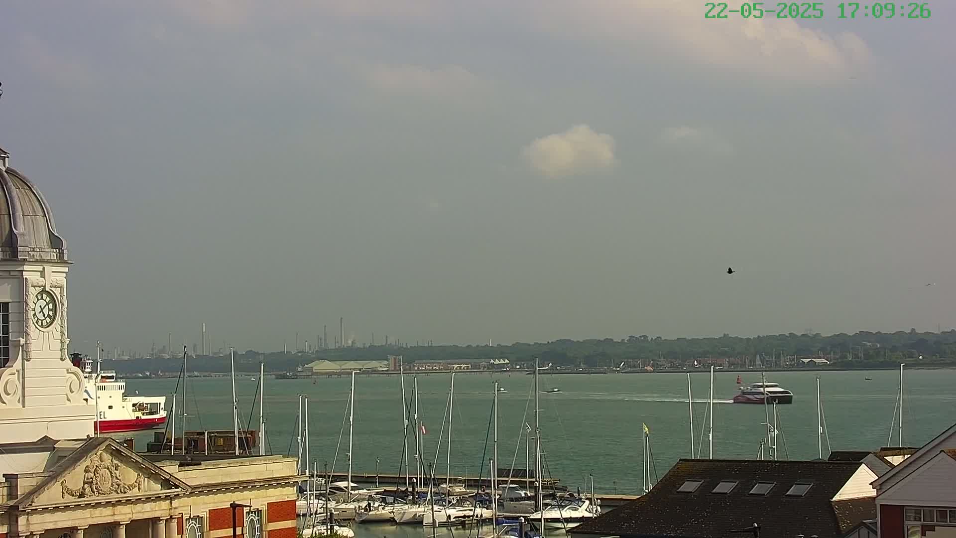 A partly cloudy day overlooks a harbor filled with sailboats, a ferry, and a building with a clock tower, with industrial structures visible in the distance.