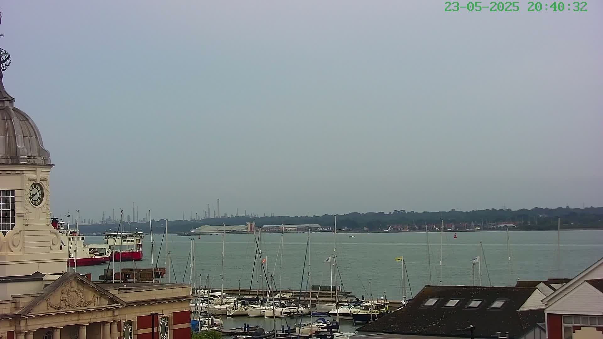A clock tower overlooks a calm harbor filled with sailboats and a few larger vessels, with a distant industrial landscape visible under a hazy sky.