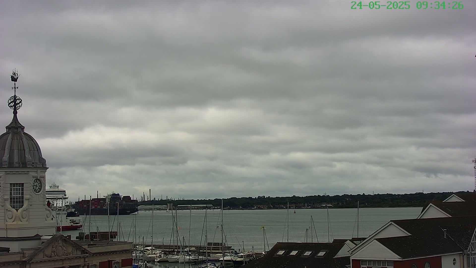 A harbor scene under a cloudy sky shows a large container ship and cruise ship in the distance, with a smaller harbor filled with sailboats closer to the viewer, and a building with a cupola in the foreground.