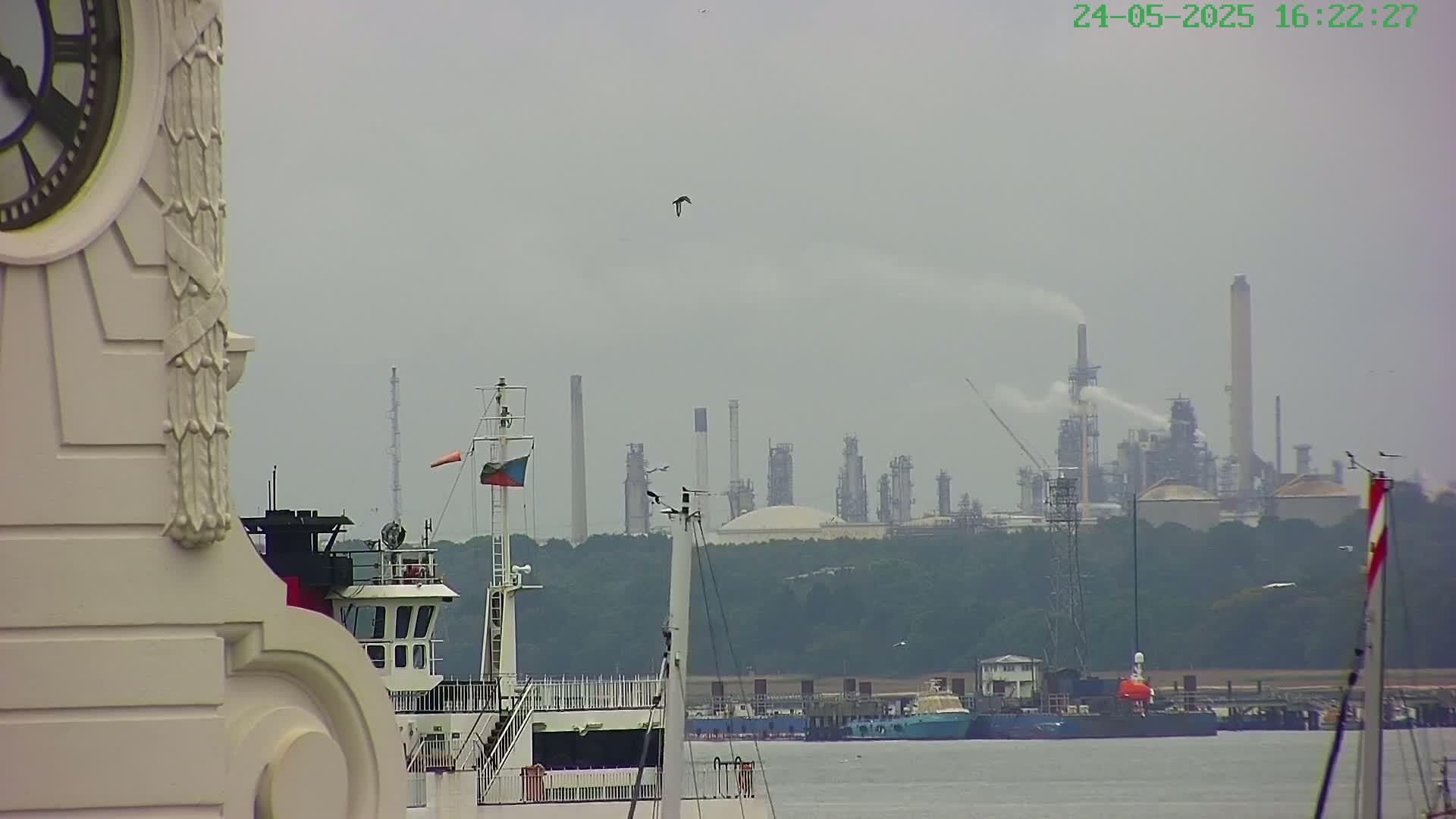 A hazy view from beside a clock tower shows several ships on calm water with an industrial refinery in the background.