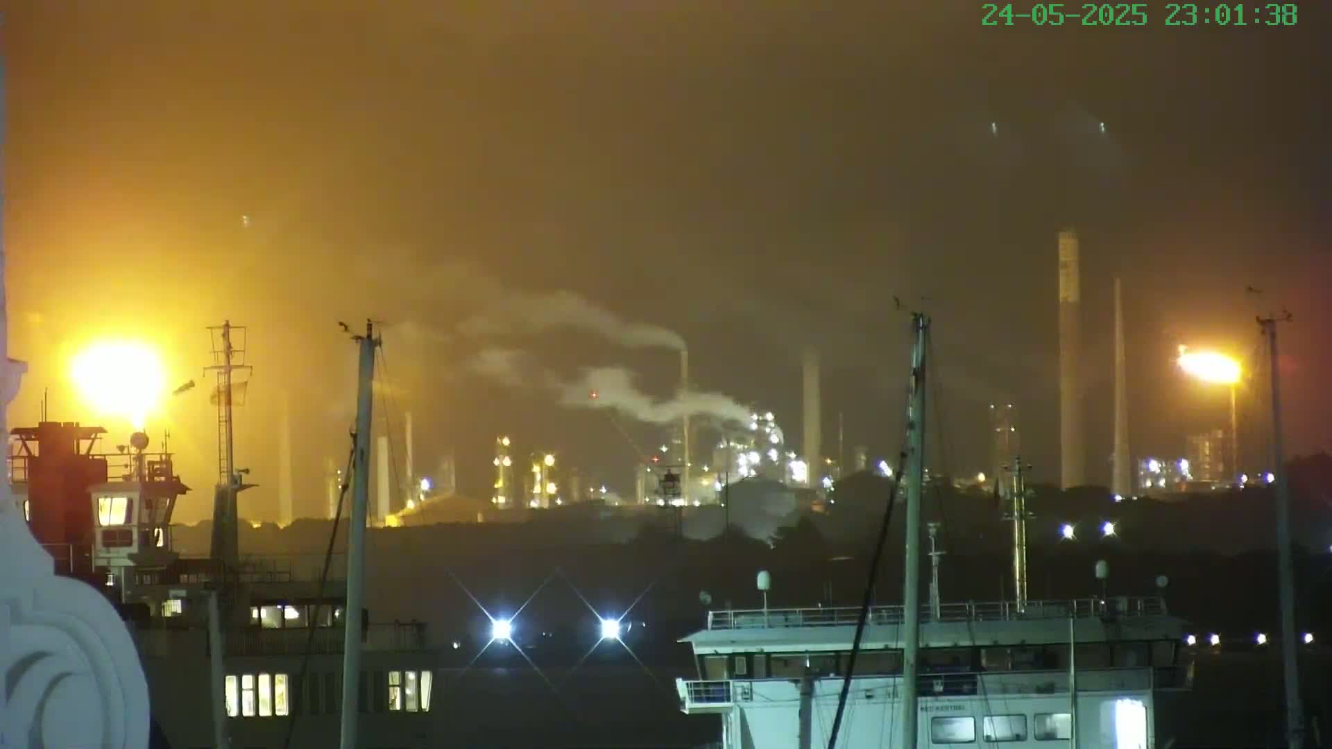 A hazy night scene shows an illuminated industrial plant emitting smoke, with ships and masts in the foreground.