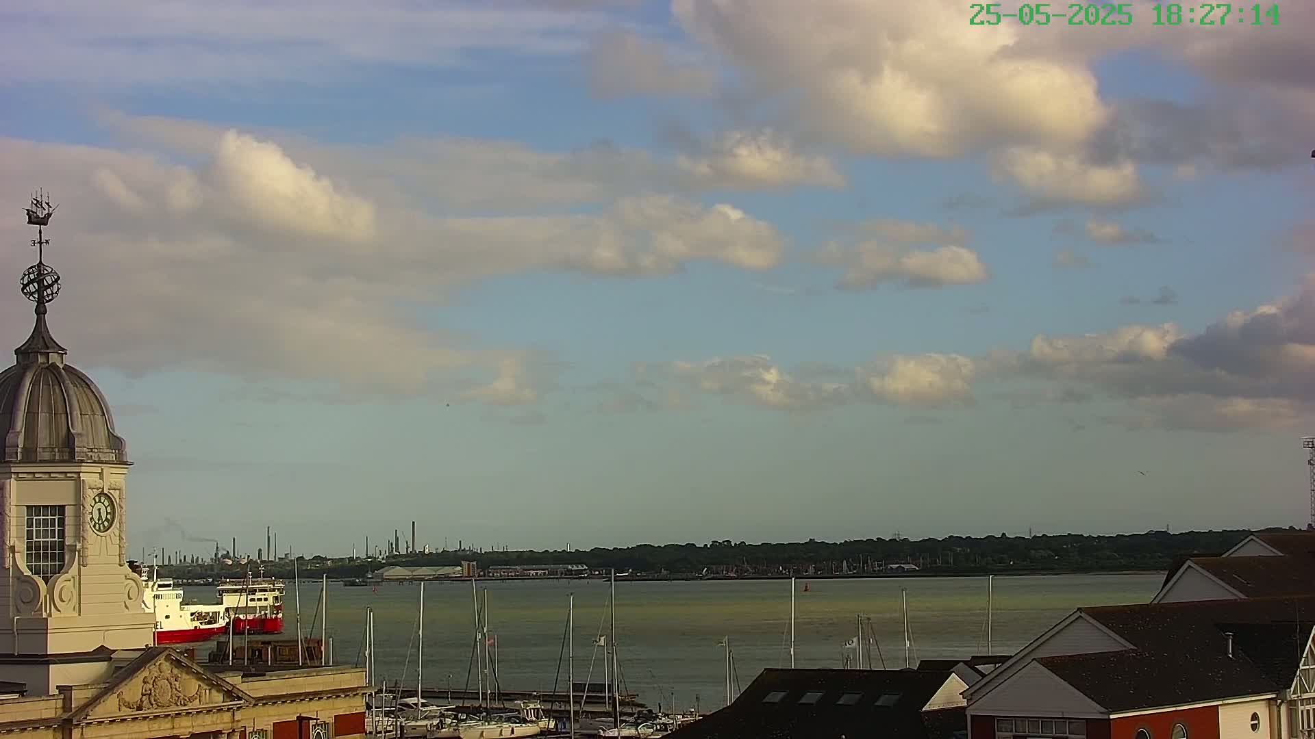 A partly cloudy sky overlooks a harbor with numerous boats and ships, a distant industrial skyline, and buildings with dark roofs.