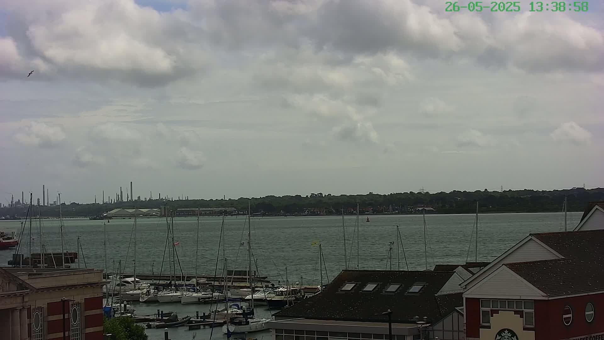 A partly cloudy day overlooks a marina filled with sailboats and a body of water leading to an industrial coastline in the distance.