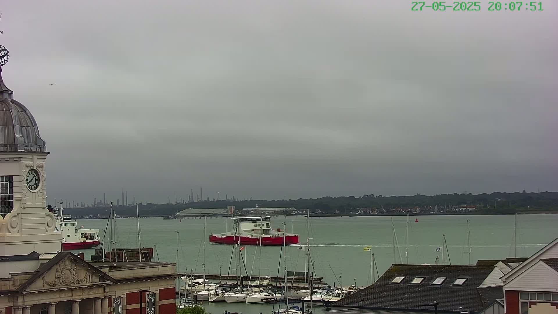 A red ferry sails past a harbor filled with sailboats under an overcast sky.