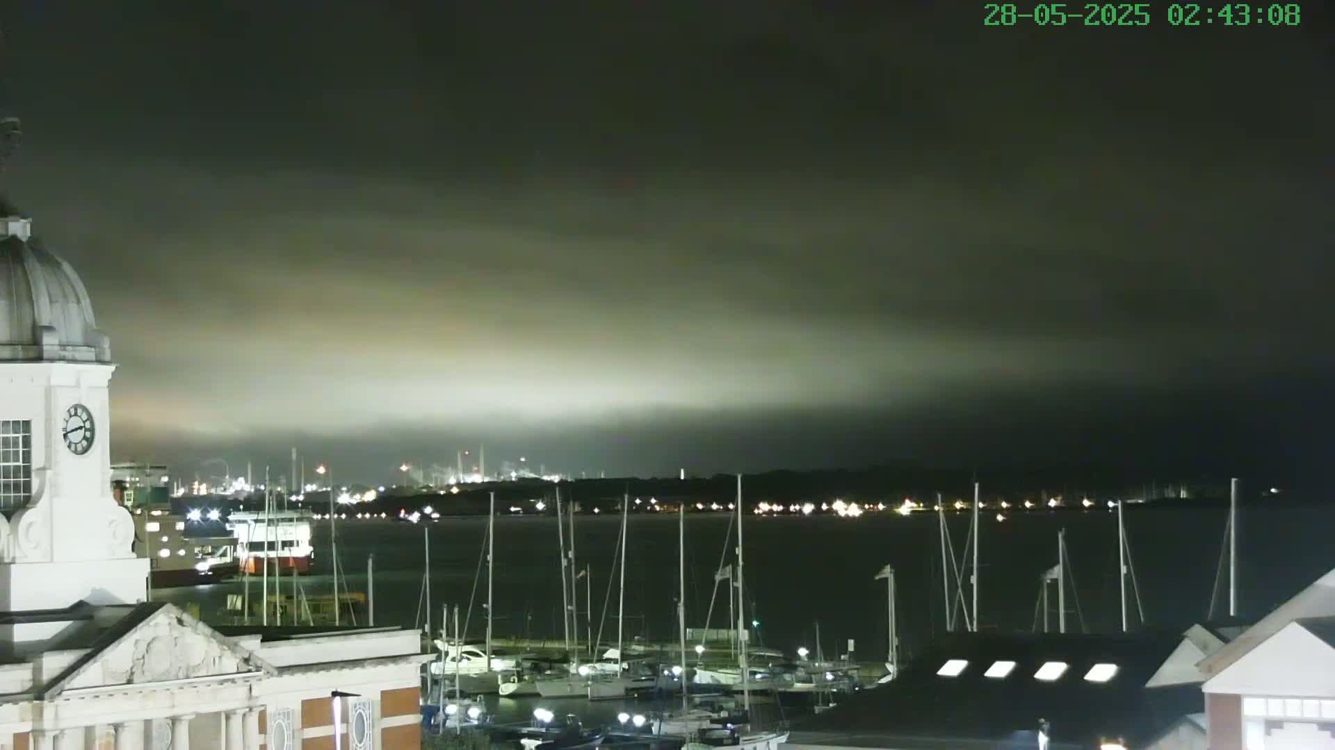 A nighttime view from a building with a clock tower shows a harbor full of boats under a dark, cloudy sky with faintly lit distant industrial areas.