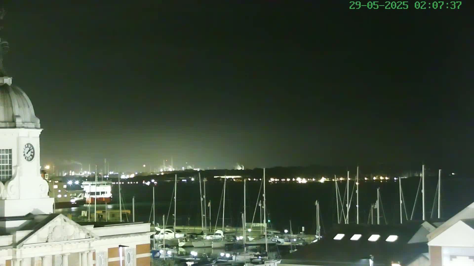 A nighttime view of a harbor filled with docked sailboats, a clock tower, and an industrial area faintly visible in the distance under a dark sky.