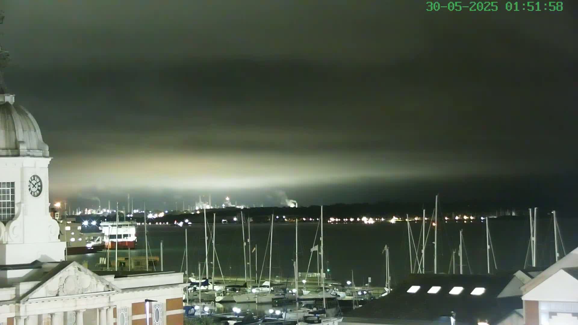 A nighttime view of a harbor filled with docked sailboats, a distant industrial area with faint lights and smoke, and a white clock tower in the foreground under an overcast sky.