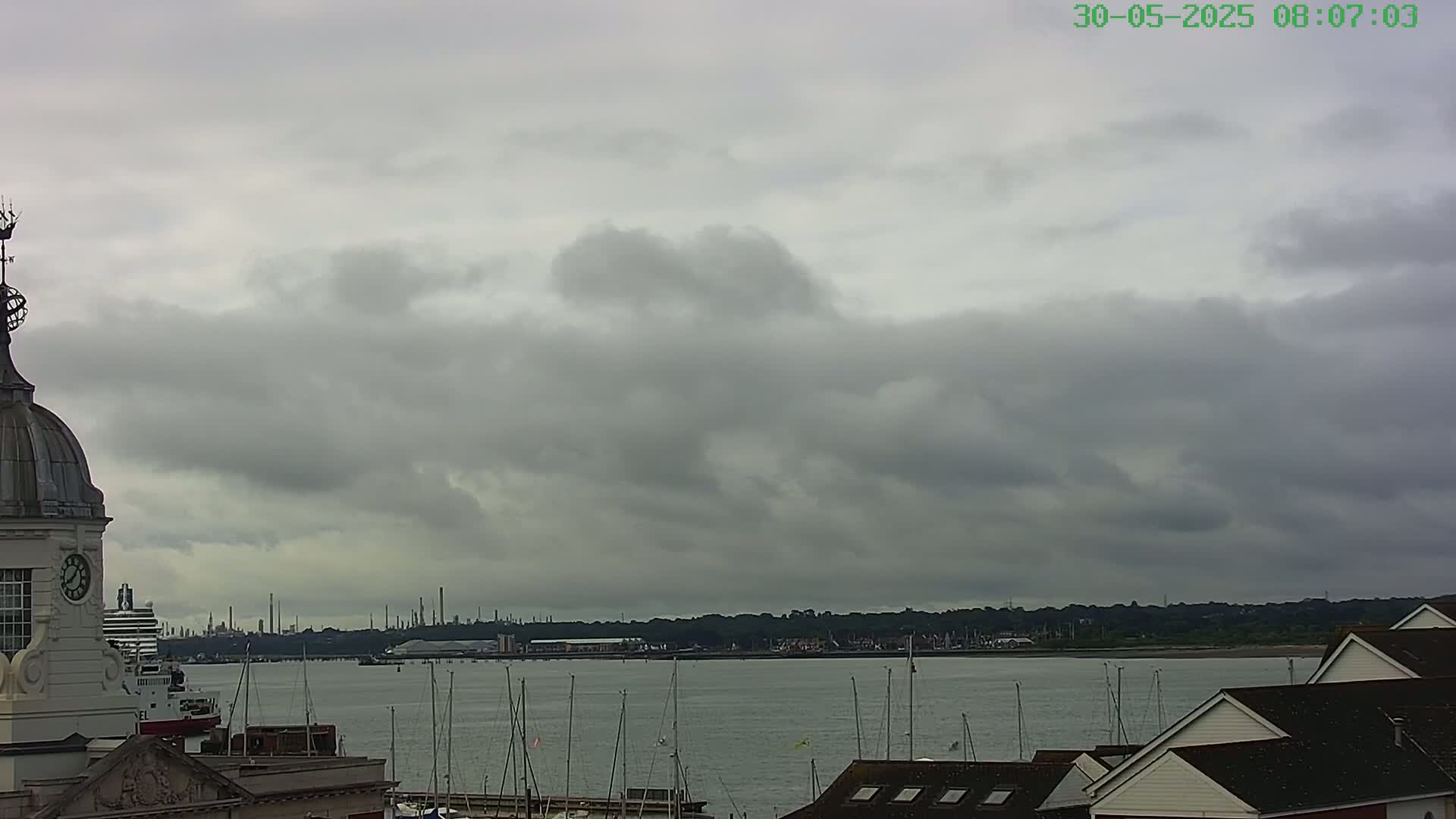 A view of a harbor with numerous boats, a distant industrial skyline, and overcast skies, seen from atop a building with a dome-shaped clock tower.