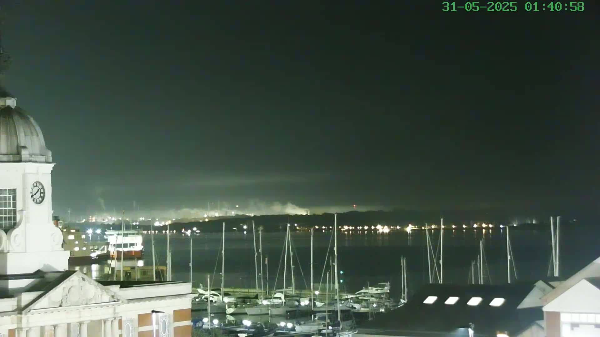 A nighttime view from atop a building shows a calm body of water with numerous sailboats docked, a distant industrial area faintly illuminated, and a hazy atmosphere.