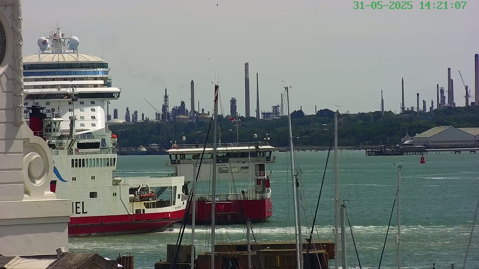 A large cruise ship and a smaller ferry are in a harbor on a partly sunny day, with an industrial plant visible in the background.