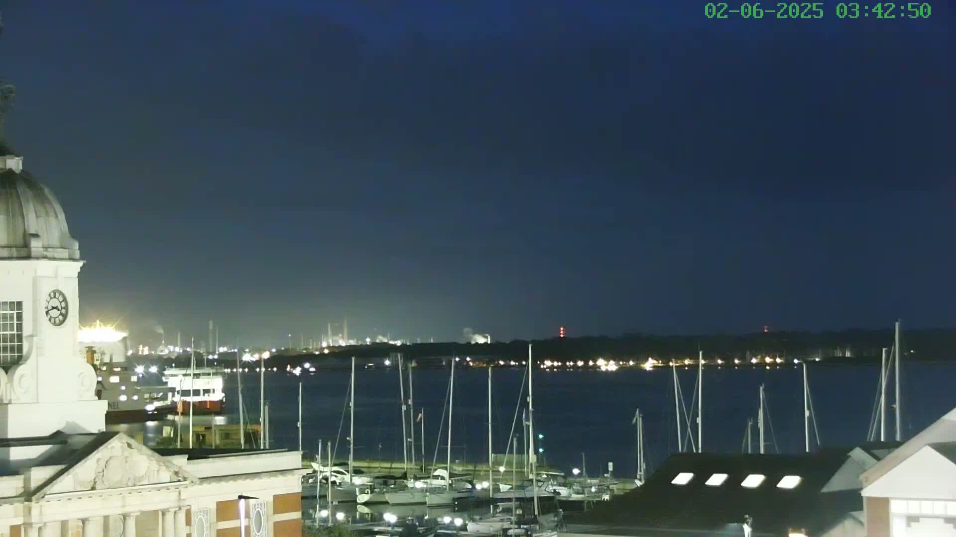 A nighttime view from atop a building shows a harbor filled with sailboats, a distant industrial area, and a dark, clear sky.