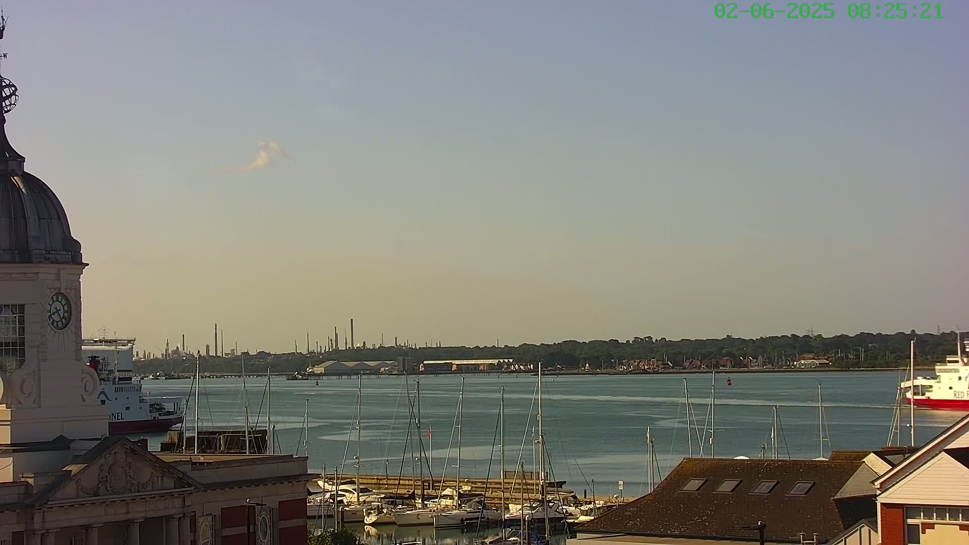 A partly cloudy day reveals a harbor scene with numerous sailboats docked, a ferry in the distance, and an industrial area visible on the horizon, all viewed from atop a building with a dome-shaped clock tower.