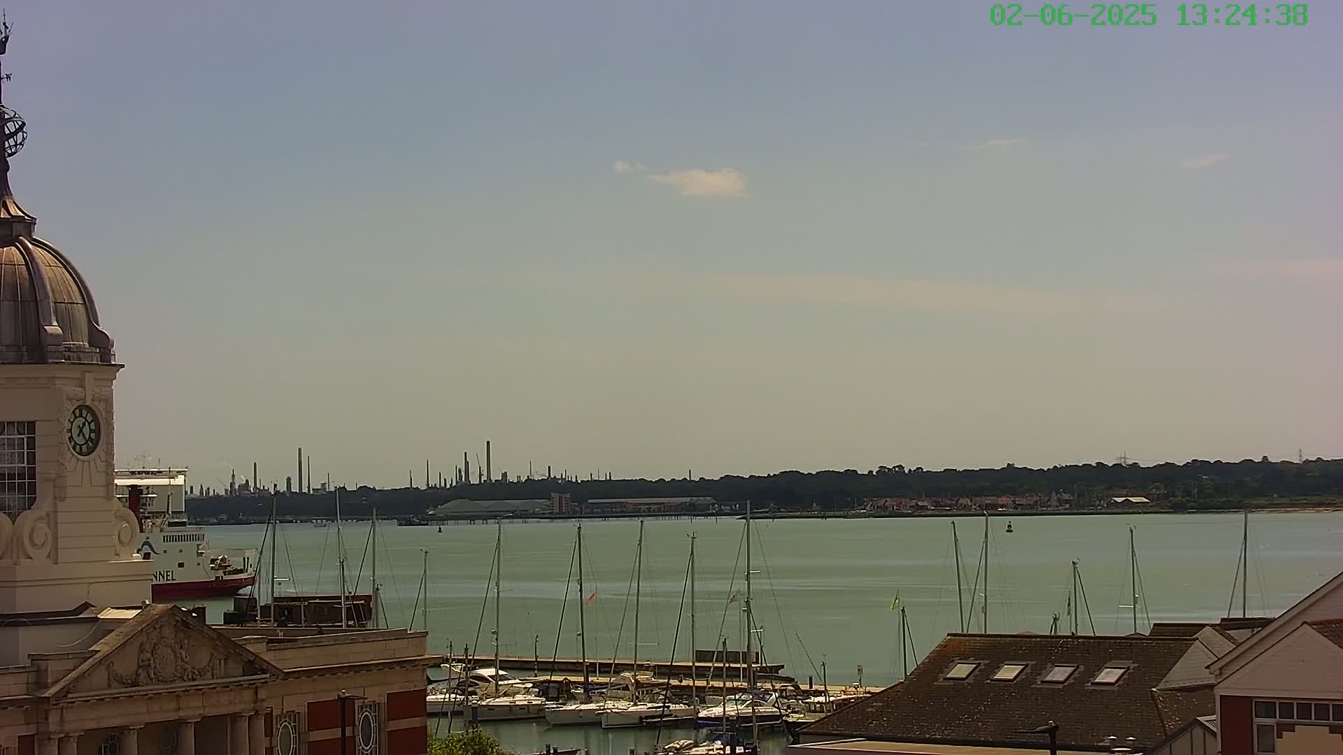 A harbor with numerous sailboats is seen from a vantage point overlooking a building with a dome and clock tower, under a mostly clear sky with a few clouds, and a distant industrial skyline is visible across the water.