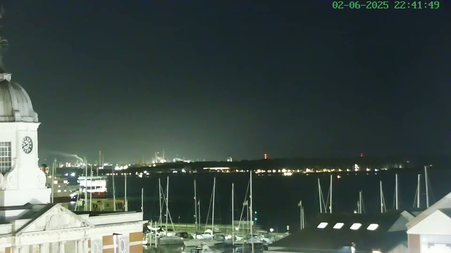 A nighttime view from atop a clock tower shows a harbor full of docked sailboats, with an industrial area faintly visible across the water under a dark sky.