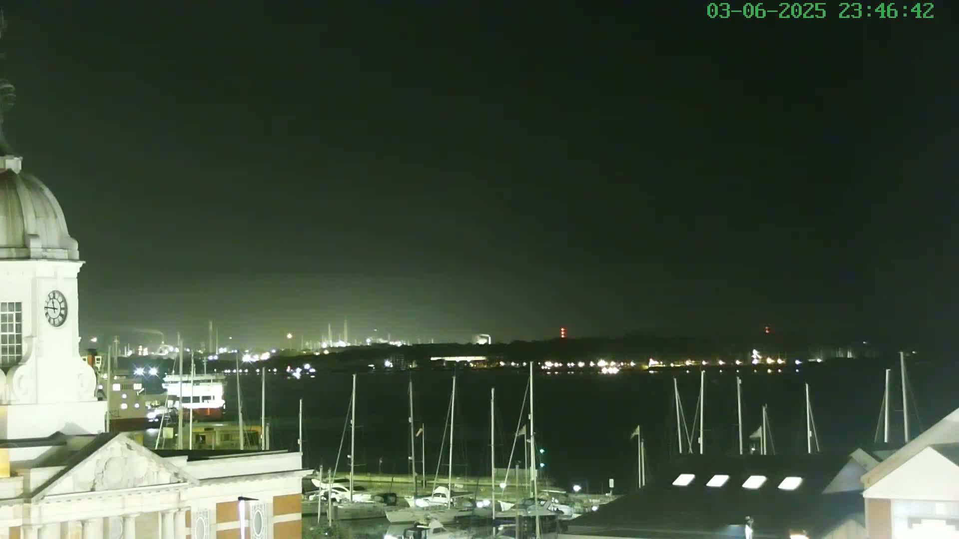 A nighttime view shows a harbor with many sailboats docked, a clock tower, and an industrial area faintly illuminated in the distance across the water.