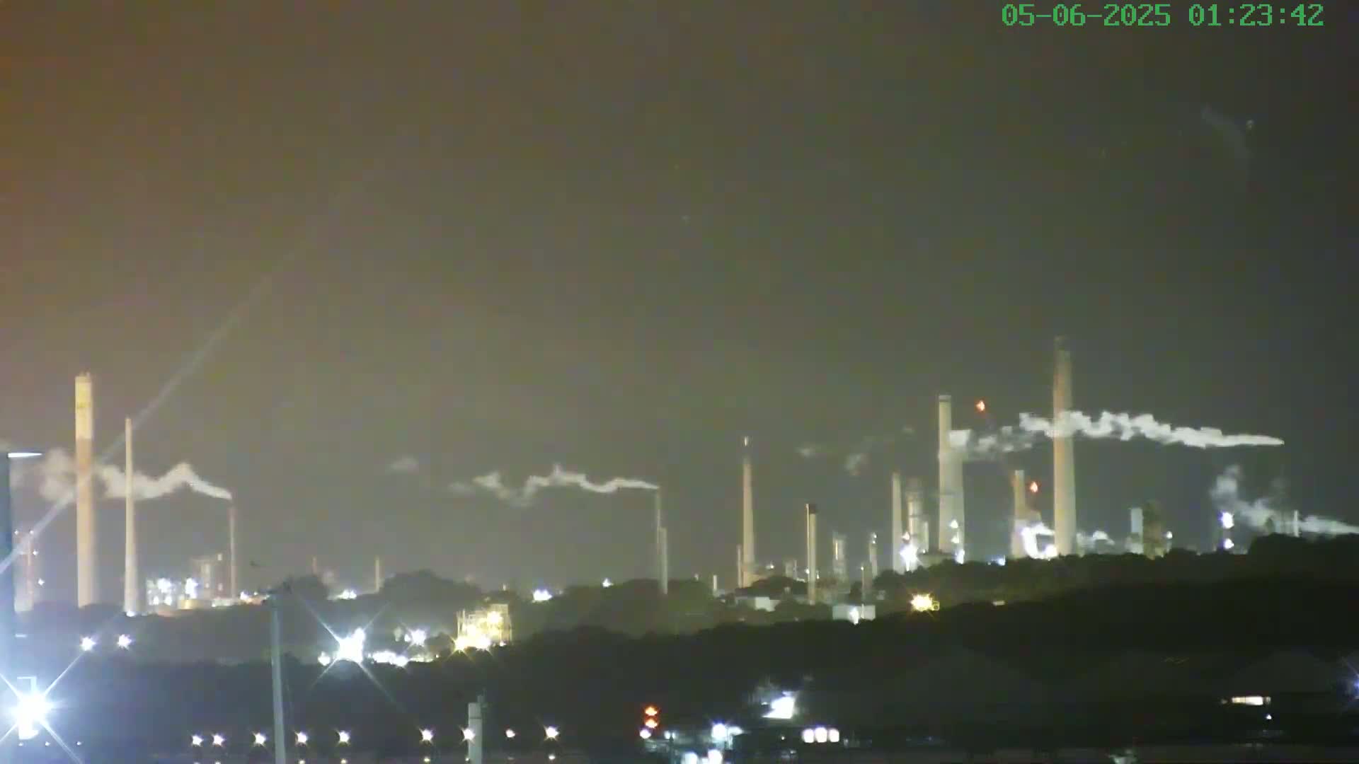 A nighttime view of an industrial plant with numerous tall smokestacks emitting plumes of smoke under a dark, hazy sky.
