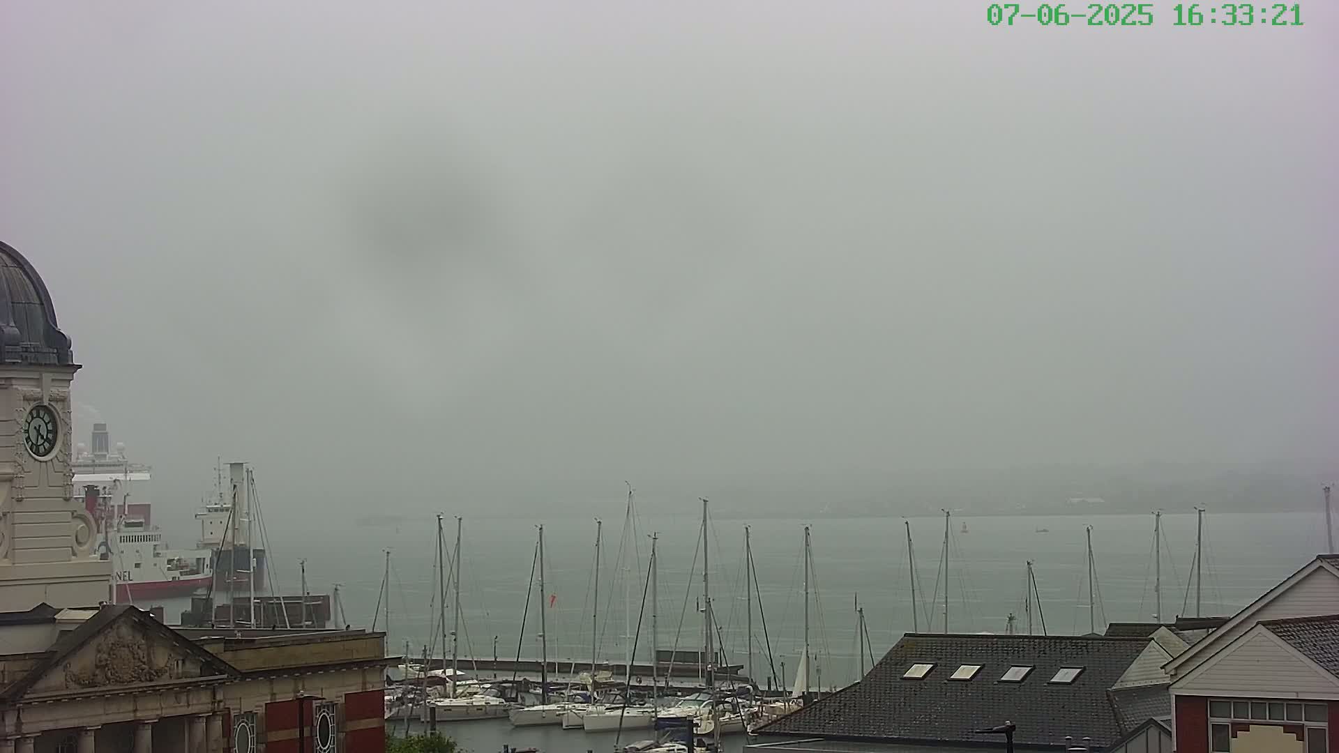 A foggy harbor scene shows a clock tower, a cargo ship, numerous sailboats moored at a marina, and several buildings.