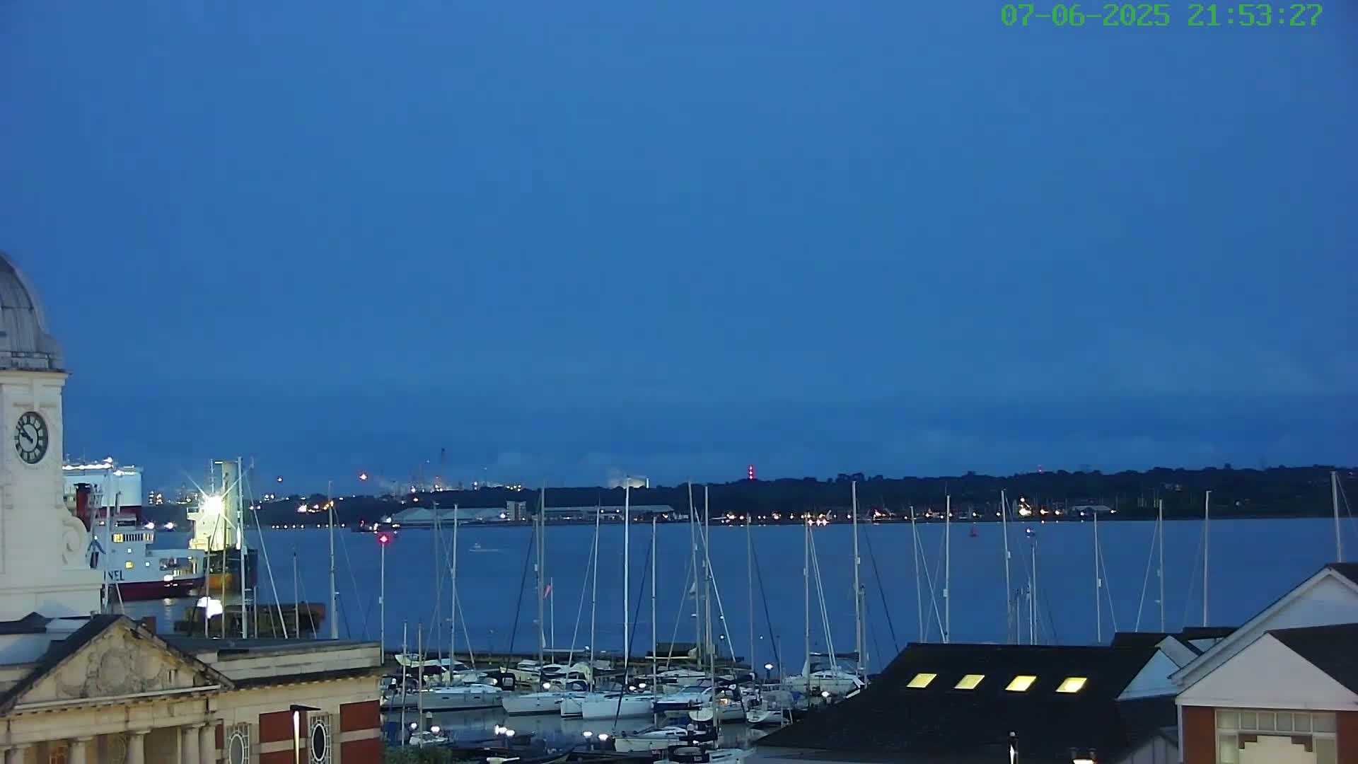 A harbor filled with sailboats is seen at twilight under a clear, dark blue sky, with a large ship and industrial buildings visible in the distance.