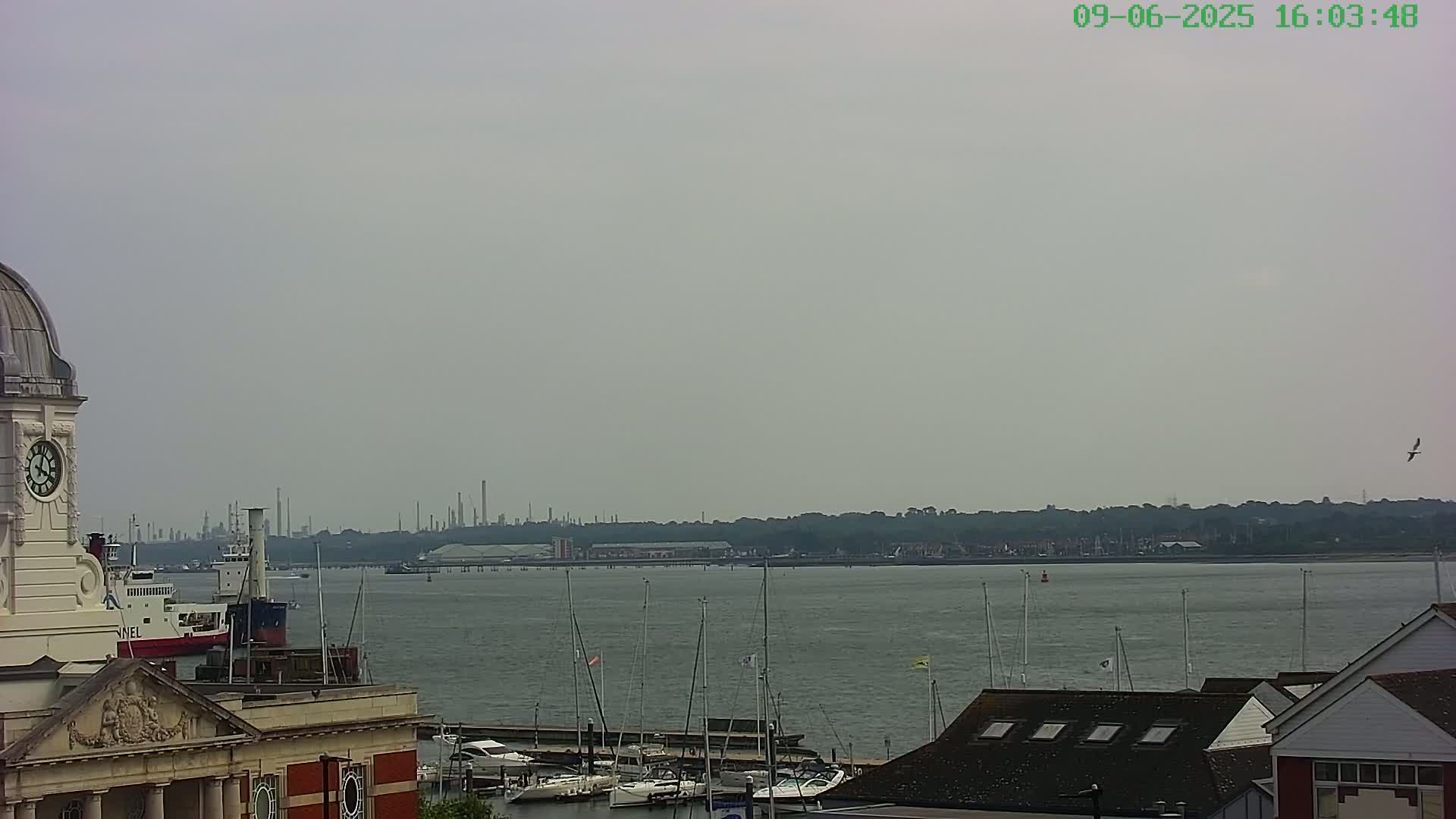 A clock tower overlooks a calm body of water with moored sailboats and a distant industrial skyline under an overcast sky.