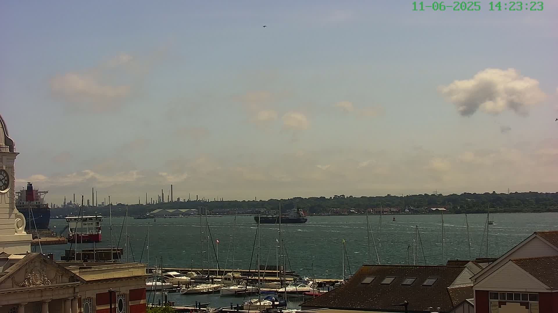 A partly cloudy day shows a harbor with many sailboats docked, a large cargo ship in the water, and an industrial area in the distance.