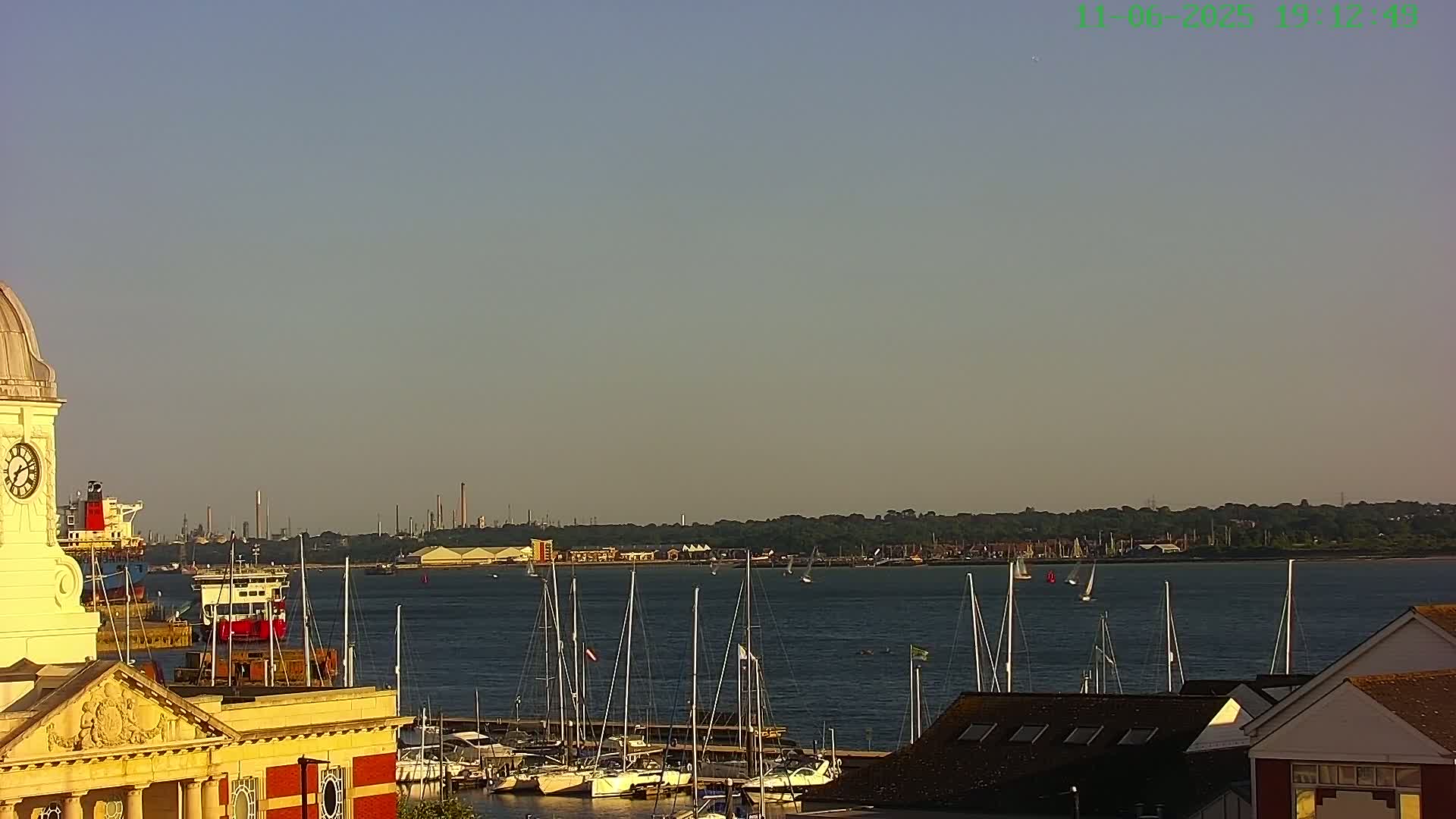 A pale yellow clock tower overlooks a calm body of water with numerous sailboats docked and a few underway, under a clear, light-blue sky.