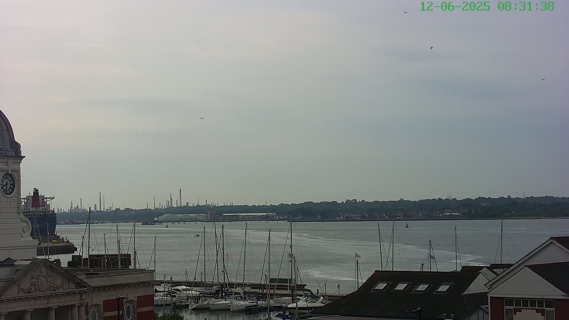 A cloudy day reveals a harbor filled with sailboats, a distant industrial skyline, and buildings with a clock tower in the foreground.