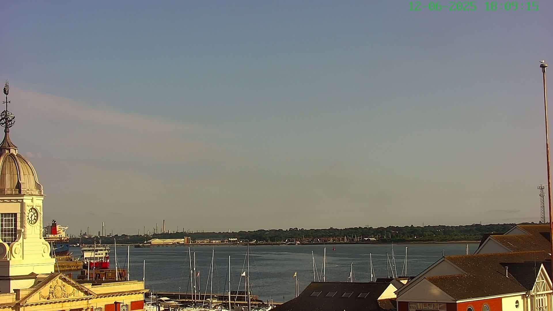A clear, sunny day reveals a harbor filled with docked sailboats, a distant industrial shoreline, and a building with a cupola and clock.