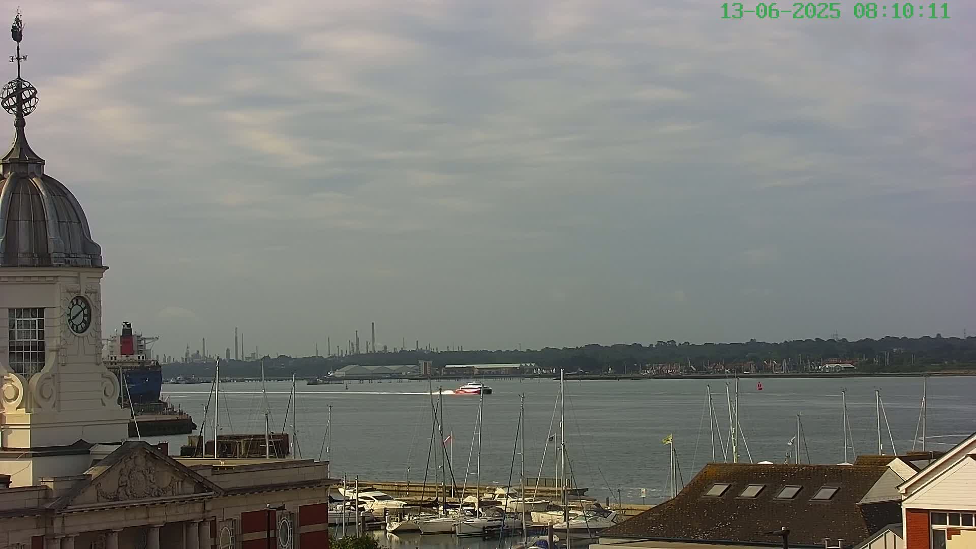 A high-angle view shows a harbor with numerous sailboats, a passing ferry, and a large building with a dome and clock tower in the foreground under a mostly cloudy sky.