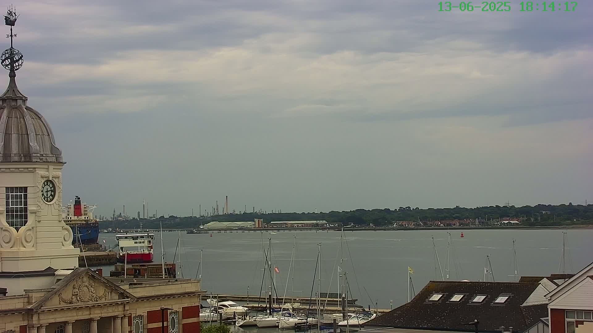 A view from atop a clock tower shows a calm body of water with sailboats docked, a distant industrial area, and an overcast sky.