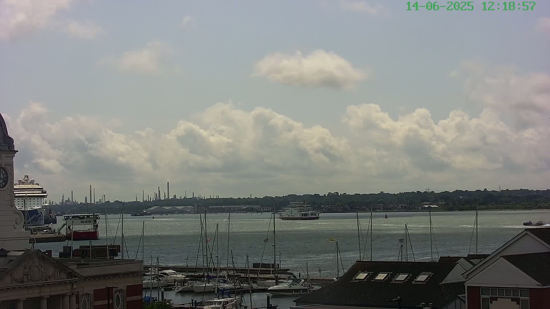 A partly cloudy sky overlooks a harbor with docked sailboats, a cruise ship, a ferry, and an industrial area in the distance.