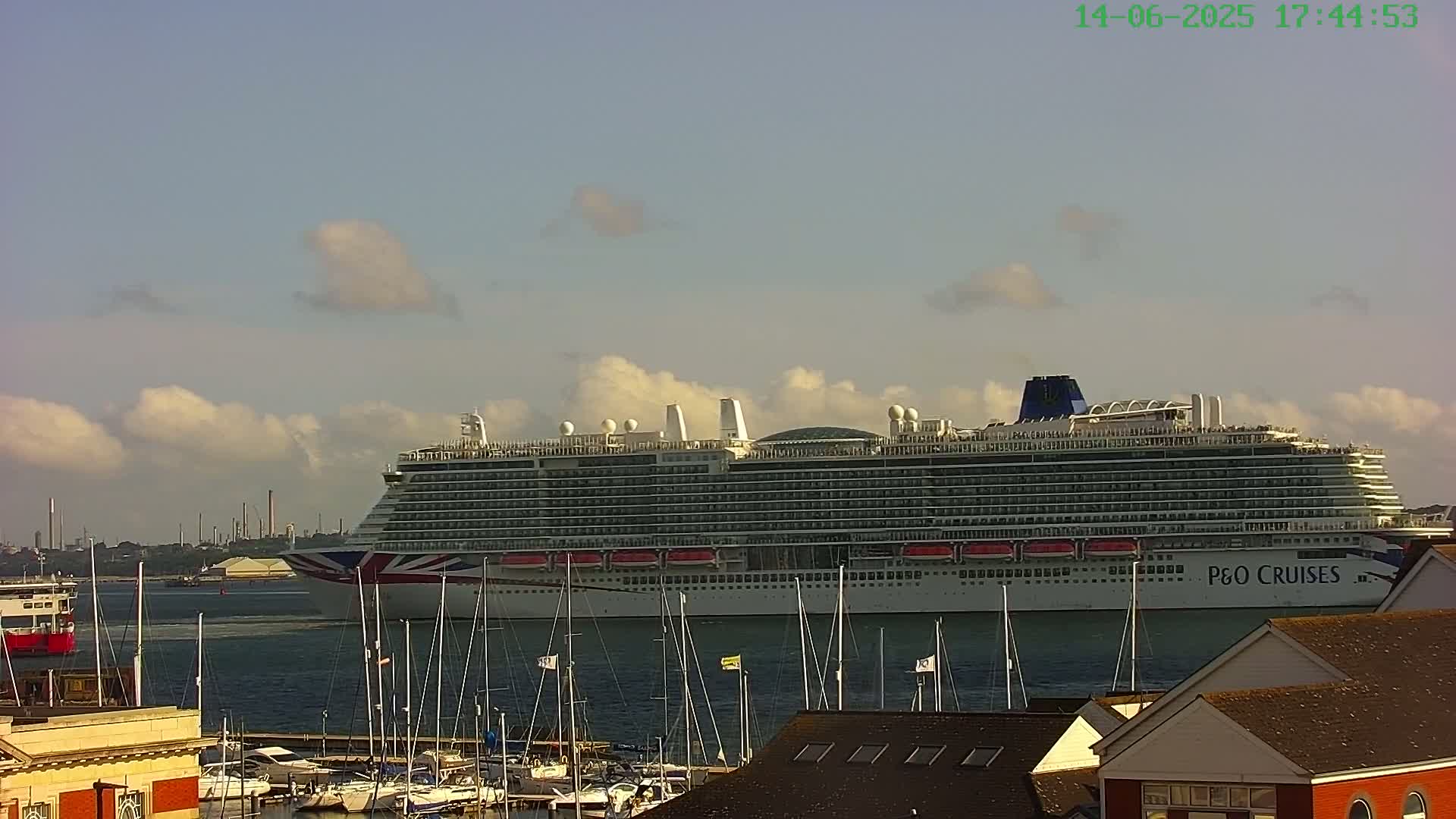 A large cruise ship is in a harbor on a partly cloudy day, with numerous sailboats docked in the foreground.