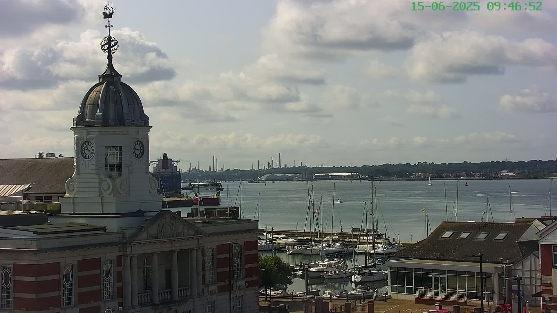 A red brick building with a white cupola and clock tower overlooks a marina filled with sailboats and yachts, under a partly cloudy sky.