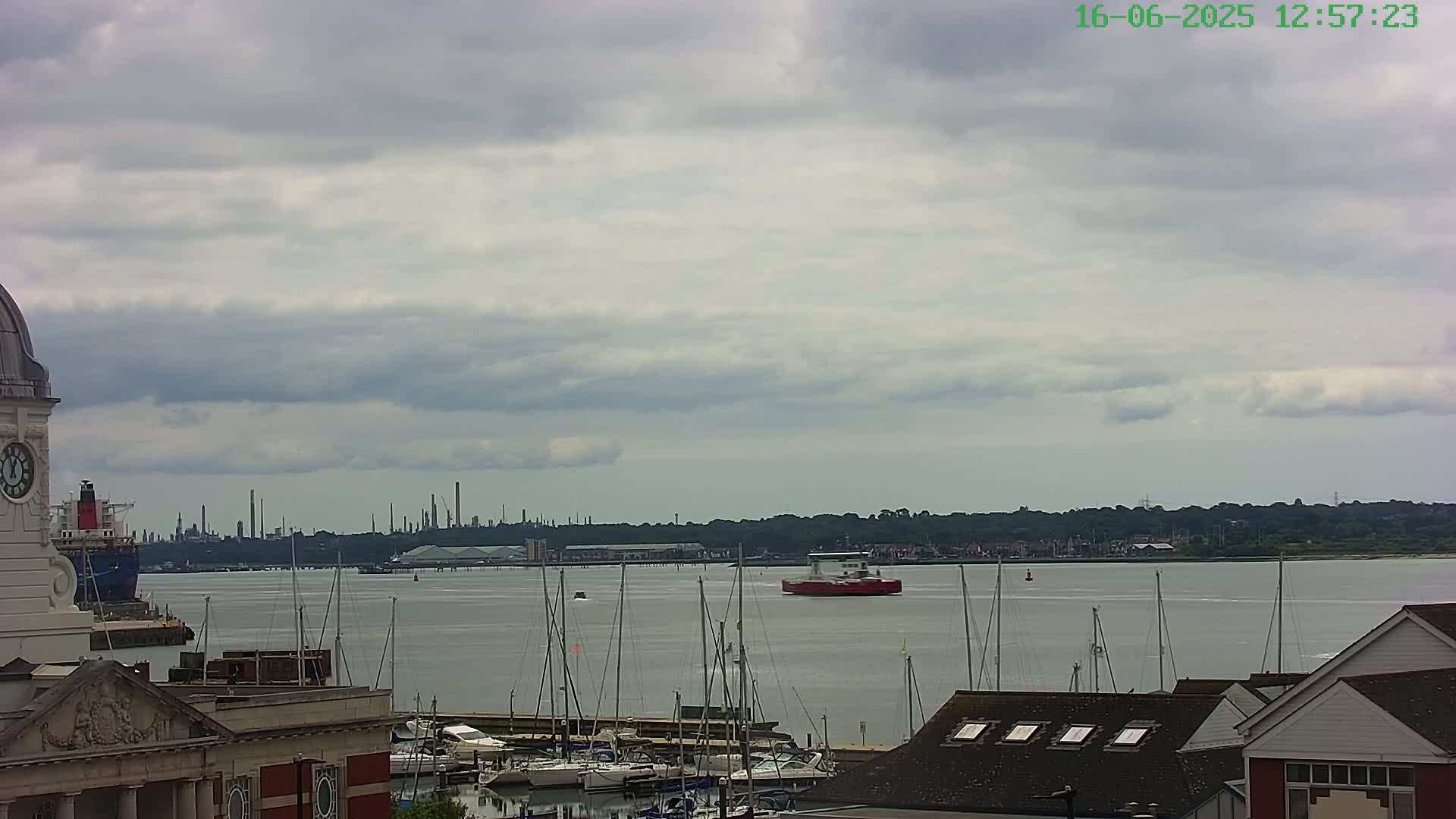 A red ferry travels across a calm body of water under a cloudy sky, with a marina full of sailboats in the foreground and an industrial skyline in the distance.