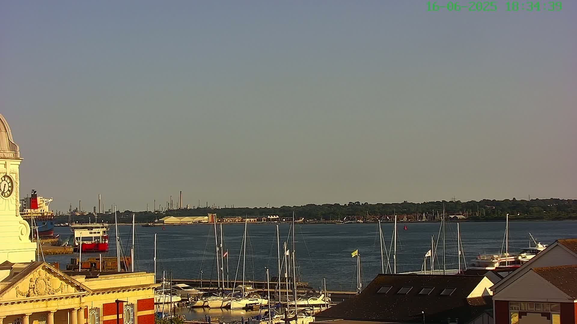A clock tower overlooks a marina filled with sailboats and motorboats, with a hazy industrial area visible across a calm body of water under a clear sky.