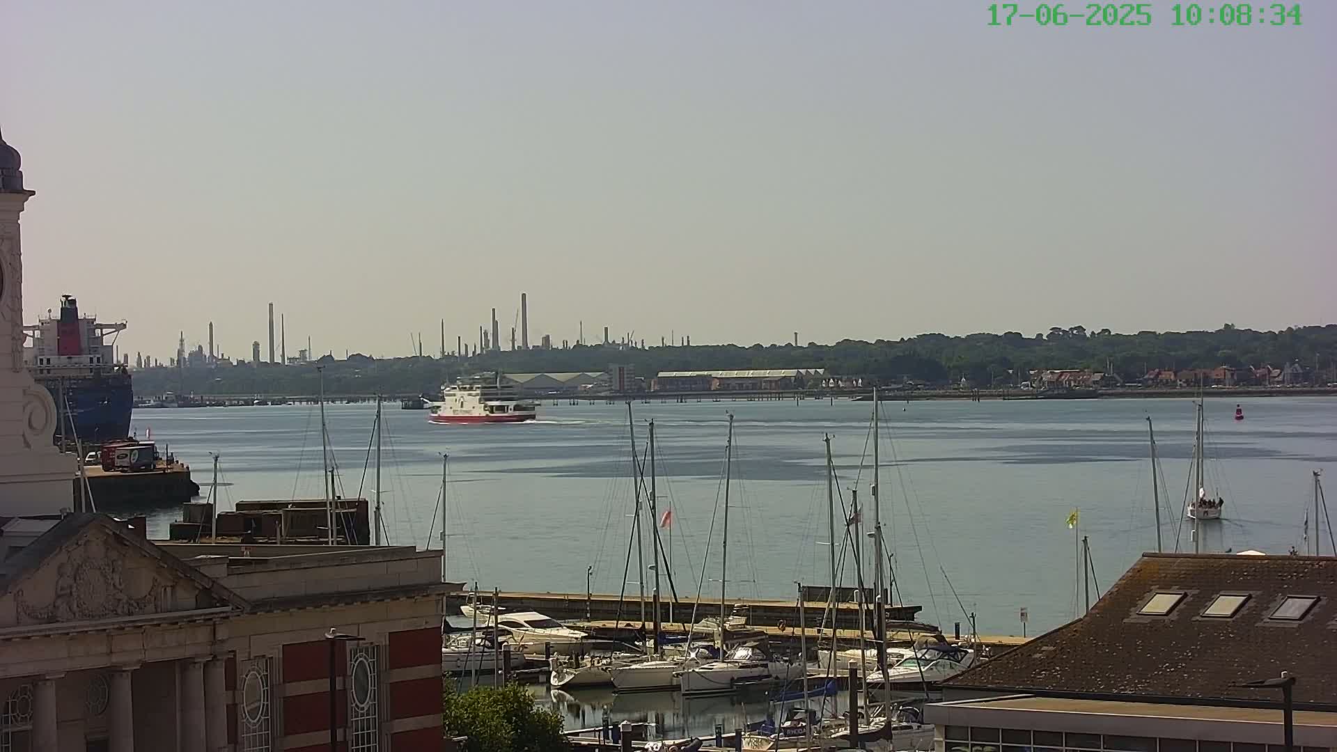 A ferry passes by a marina filled with sailboats on a hazy, sunny day, with an industrial skyline visible in the distance.