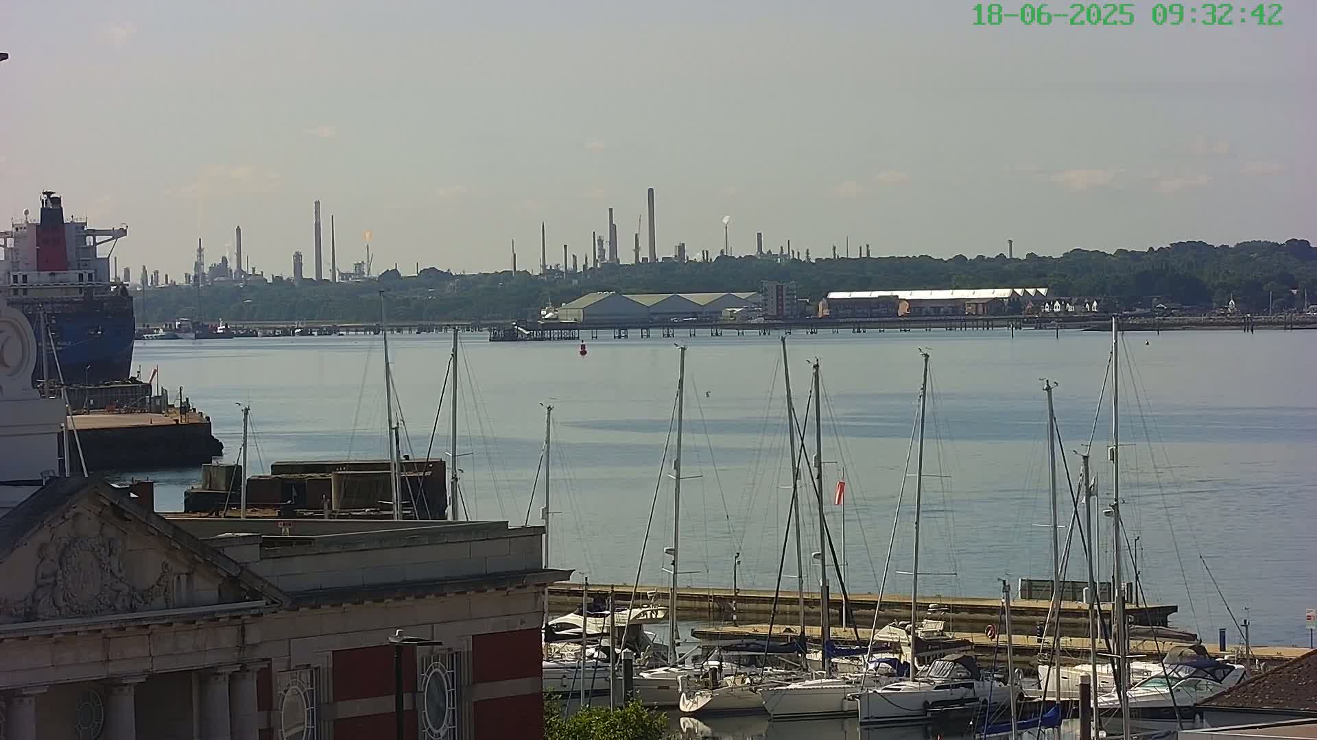 A calm body of water with numerous sailboats docked near a building, an industrial complex visible in the background under a mostly clear sky.